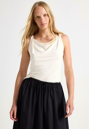 Giovane donna con lunghi capelli biondi che indossa una canotta bianca senza maniche e una gonna nera con coulisse su uno sfondo chiaro uniforme.