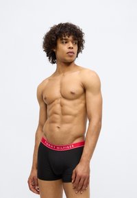 Muscular man with curly hair wearing black Tommy Hilfiger boxer briefs with a red waistband, looking to his left against a plain background.