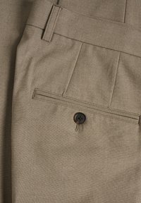 Gros plan sur un tissu beige montrant une poche arrière avec un bouton brun fixé par un fil beige sur un pantalon sur mesure.