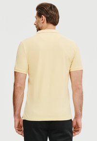 Koszulka polo w jasnym odcieniu żółtego, krótki rękaw, miękka tekstura, klasyczny kołnierzyk i boczne rozcięcia. Widok z tyłu.
