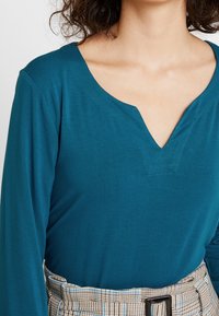 Teal langærmet top med V-udskæring og en tekstureret overflade, parret med et ternet højtaljet bælte.