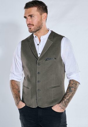 Mann mit kurzen Haaren und Tattoos, der eine graue Weste über einem weißen Hemd mit hochgekrempelten Ärmeln trägt, die Hände in den Taschen hat und nach links vor einem einfarbigen Hintergrund schaut.