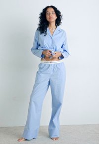 Ljust blårandig pyjamasset som innehåller en knapp-up skjorta med lång ärm och lösa byxor, med en vit märkesmidja.