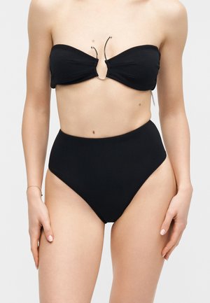 Femme portant un bas de bikini noir taille haute et un haut de bikini bandeau avec des accents en métal argenté incurvés au centre.