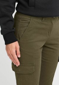 Pantaloni cargo verde oliva con una texture liscia, dotati di tasche laterali e un design aderente. La mano del modello è appoggiata sulla gamba.