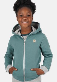 Teal zip-up hoodie med grå detaljer; har en känguruficka, mjuk textur och en logotyp på vänster bröst. Heltäckande huva.