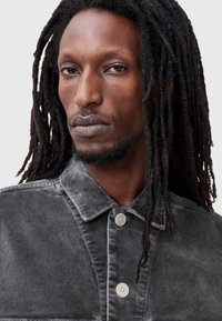 Chaqueta de mezclilla gris con cuello, que presenta un cierre de botón y una textura sutil. El tono de piel es medio con dreadlocks oscuros y largos.