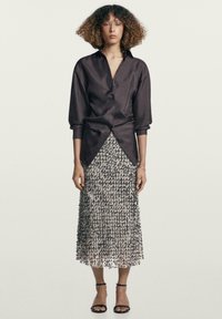 Camicia di raso scuro con bottoni abbinata a una gonna di media lunghezza in tessuto strutturato e paillettato, nei toni argento e crema, completata da sandali neri con cinturini.