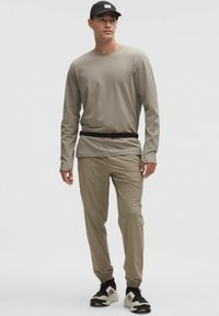 Cămașă bej cu mânecă lungă, pantaloni ușori asortați cu manșete elastice, completată de o șapcă neagră și teniși multicolori.