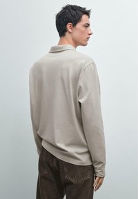 Beige poloshirt met lange mouwen en een platte kraag, zachte textuur en een relaxte pasvorm, gecombineerd met donkerbruine jeans. Eenvoudige achterkant.