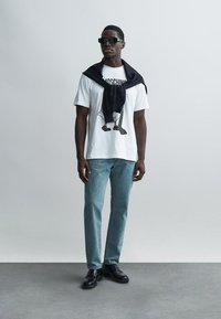 Λευκό t-shirt με γραφικό σχέδιο και μαύρο τζιν. Μαύρο πουλόβερ περασμένο στους ώμους; μαύρα γυαλιά ηλίου. Μαύρα παπούτσια.