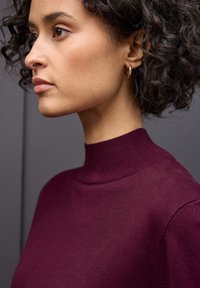 Pull à col roulé bordeaux en tricot fin, avec un col côtelé. L'arrière-plan est gris, créant un contraste avec la couleur du pull.