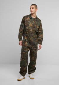 Katonai mintás overál, zöld, barna és fekete színekben, rugalmas derékkal, cipzáras zsebekkel és tartós anyagból. Fehér sportcipőkkel párosítva.