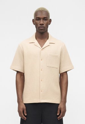 Homme aux cheveux blonds courts portant une chemise beige à manches courtes texturée avec une poche poitrine, debout devant un fond blanc uni.