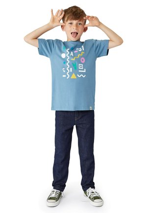 Hellblaues T-Shirt mit kurzen Ärmeln, das bunte geometrische Muster zeigt, kombiniert mit dunklen Jeans und grünen Schuhen mit weißen Schnürsenkeln.