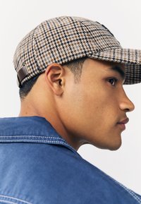 Braun-beige Hahnentritt-Mütze mit einem glatten Lederband. Das Design umfasst eine strukturierte Krempe und ein strukturiertes Gewebe. Blaue Jeansjacke sichtbar.