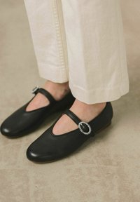 Zapatos Mary Jane de cuero negro con punta redonda, que presentan una hebilla metálica decorativa en la correa. Llevados con pantalones de color claro.