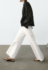 Black lightweight long-sleeve top with loose fit and pleated texture, paired with white wide-leg trousers and flat sandals.