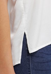 Weiße Bluse mit kurzen Ärmeln und seitlichem Schlitz, aus einem weichen, glatten Stoff gefertigt. Mit gesäumten Kanten und modernem, lockerer Passform. Über dunklen Jeans getragen.