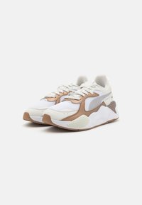 Chaussures de sport blanches et marron en mesh et matériaux synthétiques, avec bout arrondi, semelle texturée et fermeture à lacets.