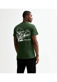 Grön bomullströja med en rygggrafik av vita blommor och text. Kortärmad och rundhalsad design, avslappnad passform och slät textur.