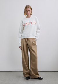 Vit sweatshirt med "SCALPERS" i orange tryck, matchad med beige vida byxor och svarta skor. Enkel design, avslappnat tyg.