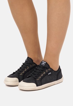 Baskets en toile noire avec embouts et semelles en caoutchouc blanc, lacets noirs, portées pieds nus sur un fond uni.