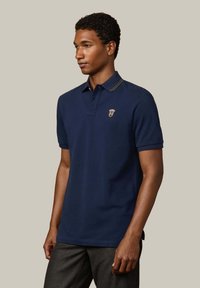 Granatowa koszulka polo z bawełny, z dwiema guzikami na stójce, żebrowanym kołnierzykiem z zielonymi detalami oraz małym haftowanym logo na piersi.