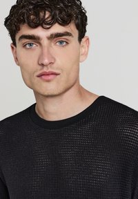 Giovane uomo con capelli ricci scuri e occhi blu che indossa un maglione a maglia nera con texture su uno sfondo chiaro e semplice.