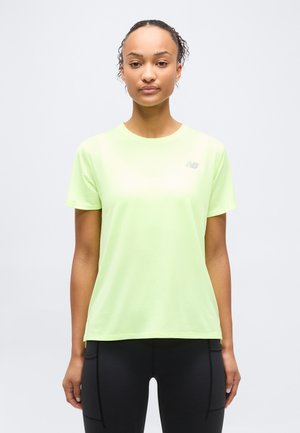 Mujer de pie, mirando al frente, vestida con una camiseta deportiva de manga corta color verde claro y leggings negros, frente a un fondo liso.