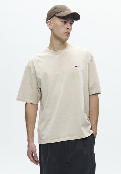 Jeune homme portant un t-shirt beige oversize avec un petit logo "W.W.", un pantalon foncé et une casquette marron, regardant sur le côté avec les mains détendues.