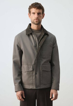 Hombre que lleva una chaqueta oliva con grandes bolsillos delanteros y un cuello oscuro, de pie frente a un fondo claro y liso.