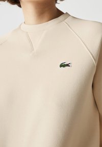 Sudadera beige con acabado texturizado, que presenta un pequeño logotipo de cocodrilo bordado en verde y blanco en el área del pecho izquierdo.