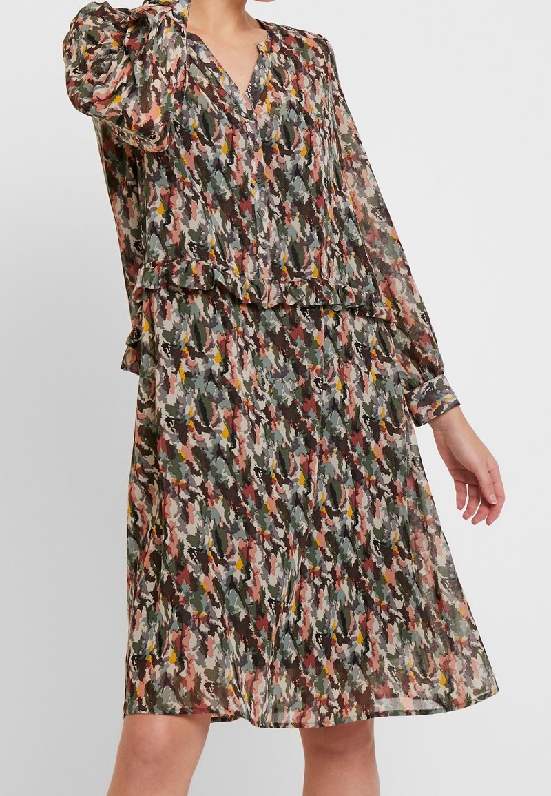 Femme portant une robe à manches longues, boutonnée devant, avec un motif abstrait multicolore et un détail volanté à la taille.