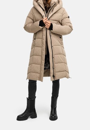 Personne portant un long manteau matelassé beige avec un grand col, un pantalon noir ajusté et des bottines noires épaisses, debout sur un fond blanc.