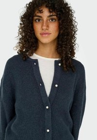 Cardigan bleu marine foncé en tissu tricoté texturé, avec un col en V, des boutons ressemblant à des perles et des manches longues. Porté par-dessus un haut léger.
