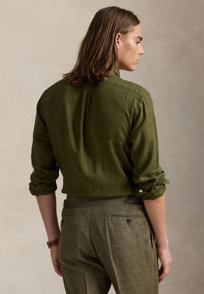 Ľanová košeľa olivovo zelenej farby s ohrnutými rukávmi, disponujúca manžetou na gombík a jednoduchým golierom, v páre s hnedými károvanými nohavicami so vzorom.