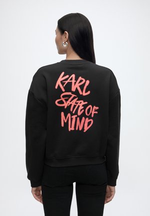 Naisel pikkade tumedate juustega, kes kannab hõbedasi rõngakujulisi kõrvarõngaid ja musta dressipluusi roosa grafititekstiga "KARL STATE OF MIND" seljal.