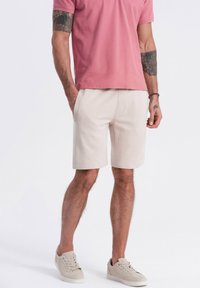 Shorts en coton beige clair avec poches latérales, associés à un t-shirt rose poussiéreux. Le modèle porte des baskets gris clair. Pas de motifs ni de détails métalliques.