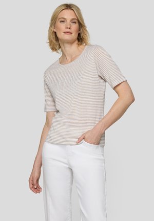 Woman with short blonde hair wearing a beige and white striped short-sleeve top and white pants, standing with one hand in pocket.