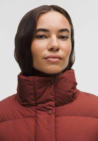 Rød polstret jakke med høj krave, der har trykknapper til lukning. Glat, blank overflade med quiltet design.
