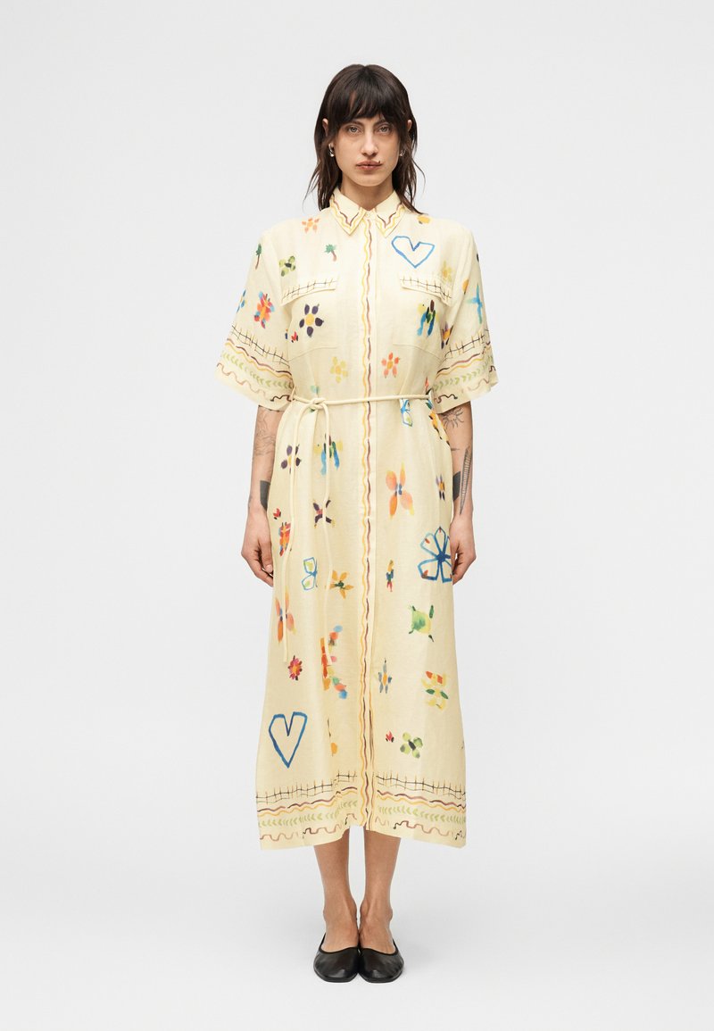 Γυναίκα που φοράει μια κρέμ μίντι φορέμα με πολύχρωμα αφηρημένα λουλουδάτα και καρδιές μοτίβα, ζώνη στη μέση, κοντά μανίκια και μαύρα επίπεδα παπούτσια.