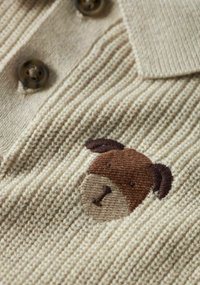 Polo beige lavorata a maglia con trama a coste, caratterizzata da un viso di cane marrone ricamato sul lato sinistro e due bottoni scuri al collo.