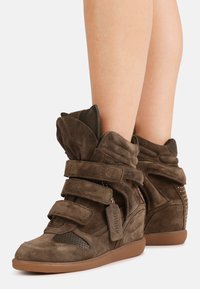 Καφέ καστόρινες σνίκερ με πλατφόρμα, διπλά λουράκια με βέλκρο, φερμουάρ στα πλαϊνά και ταμπελάκι μπρελόκ σε αντίστοιχο χρώμα, φορεμένα σε  γυμνά πόδια.