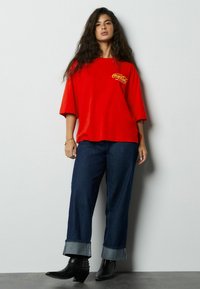 Czerwona, oversize'owa koszulka z logo Coca-Cola, zestawiona z ciemnymi, szerokimi spodniami denimowymi z podwiniętymi nogawkami oraz czarnymi, spiczastymi botkami za kostkę.