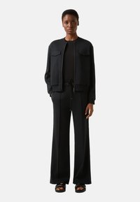 Zwarte trui met twee borstzakken, ritssluiting en geribbelde manchetten, gecombineerd met zwarte broek met brede pijpen en zwarte sandalen. Soepele textuur.