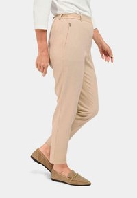 Pantalons beiges sur mesure avec une coupe slim, taille élastique, fermeture éclair sur le côté et tissu lisse. Associés à des mocassins en daim beige avec un accent doré.