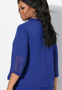 Top in tessuto azzurro trasparente con maniche a tre quarti, design aperto sul davanti e texture morbida. Presenta un orlo dritto e una leggera lucentezza.