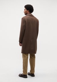 Cappotto in misto lana marrone con colletto a scollatura, maniche lunghe e vestibilità dritta, abbinato a pantaloni in corduroy beige e scarpe scure.