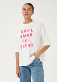 Camiseta blanca de gran tamaño hecha de algodón, con un texto rosa en negrita que dice "MORE AMOR POR FAVOR". Diseño simple con mangas cortas.